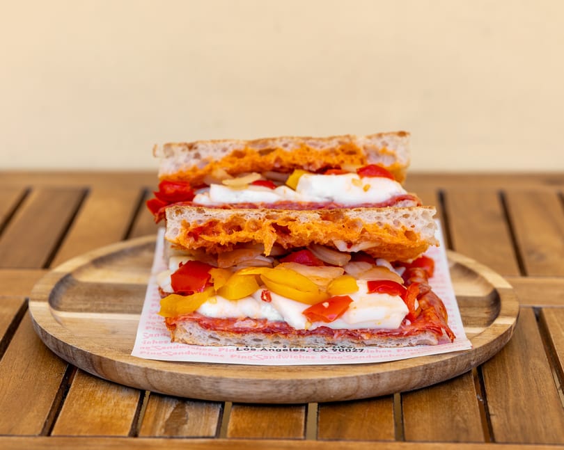 A Pino's sandwich, cut in half, filled with salami, mozzarella, peppers, and onions on a wooden plate.