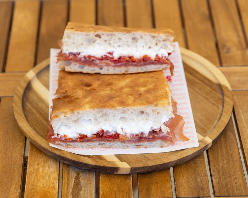 Two halves of a focaccia sandwich with prosciutto, ricotta, and roasted red peppers on a wooden plate.