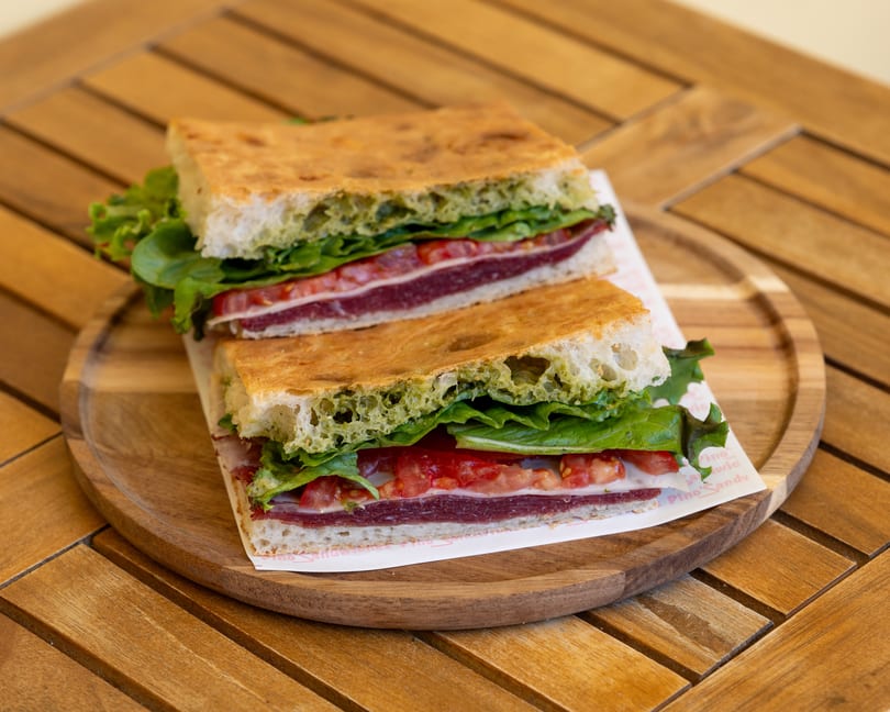 Two halves of a focaccia sandwich with pesto, lettuce, tomatoes, and meat on a wooden plate.