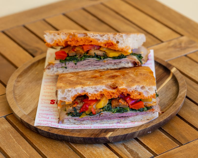 A sliced sandwich with meat, cheese, spinach, and roasted peppers on focaccia bread, on a wooden plate.