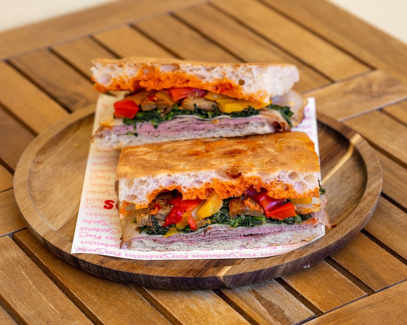 Two halves of a focaccia sandwich filled with meat, cheese, spinach, and roasted peppers on a wooden tray.