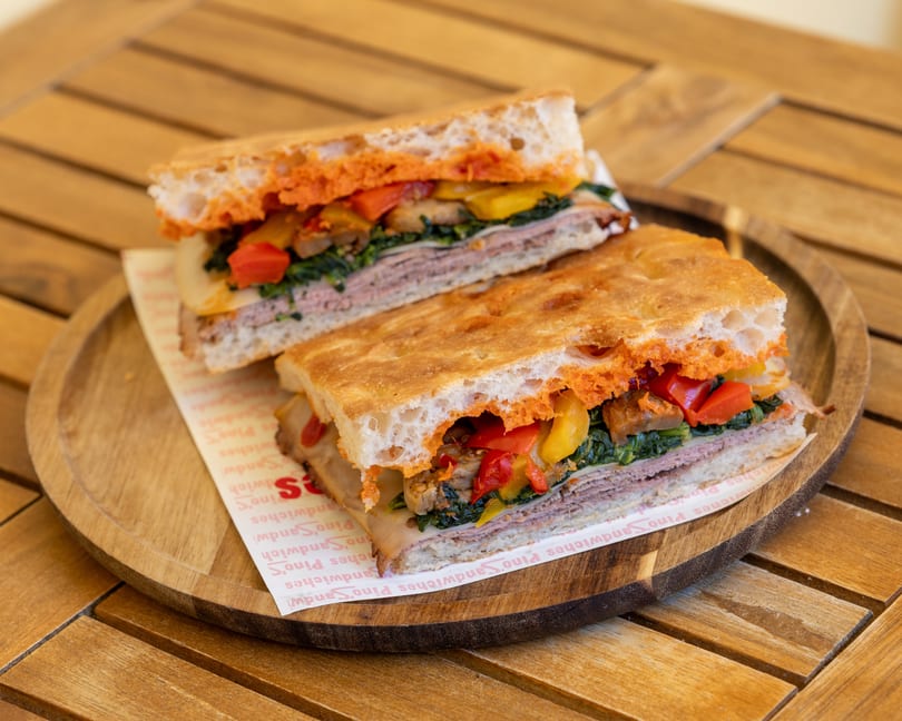 Two halves of a focaccia sandwich with meat, cheese, spinach, and roasted peppers on a wooden tray.