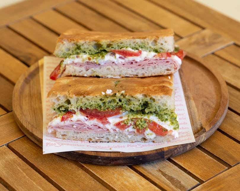 Two halves of a focaccia sandwich with ham, cheese, tomatoes, and pesto on a wooden tray.