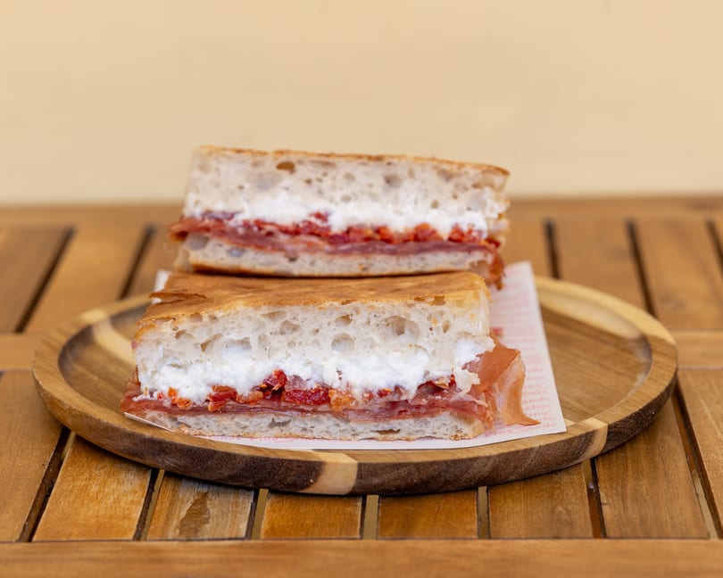 Two halves of a focaccia sandwich with prosciutto, ricotta, and sun-dried tomatoes on a wooden plate.