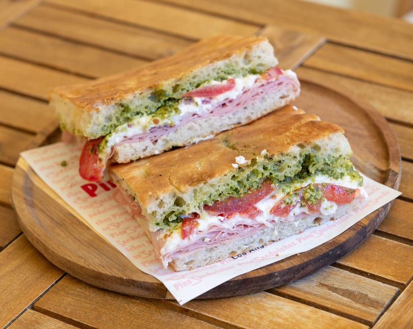 Two halves of a focaccia sandwich with ham, cheese, tomatoes, and pesto on a wooden tray.
