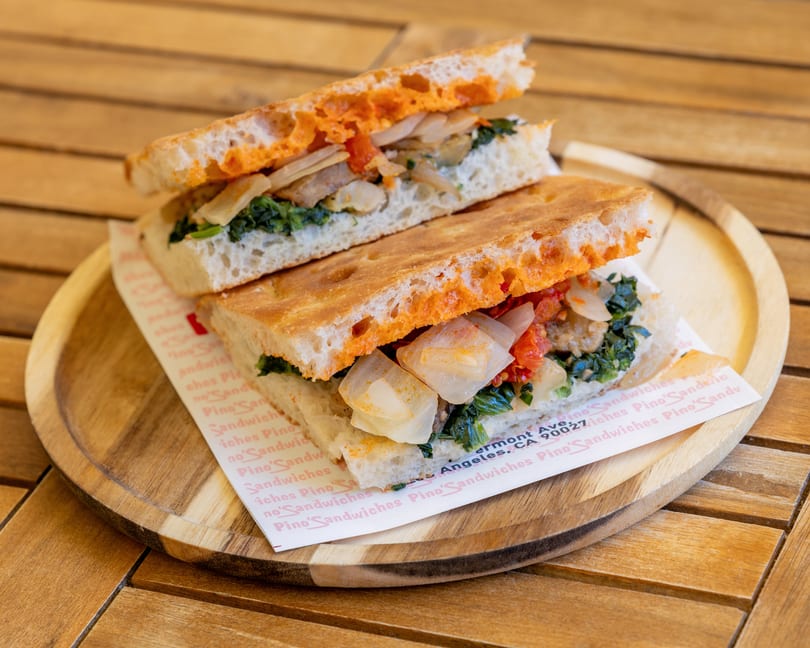 Two halves of a focaccia sandwich filled with greens, onions, and red sauce on a wooden plate.