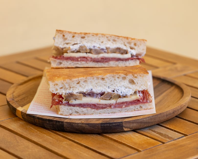 Two focaccia sandwiches with salami, cheese, and mushrooms on a wooden plate.