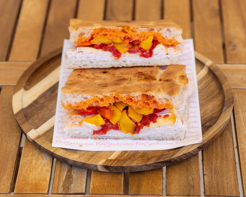 Two focaccia sandwiches with turkey, cheese, red and yellow peppers, and a red spread on a wooden tray.