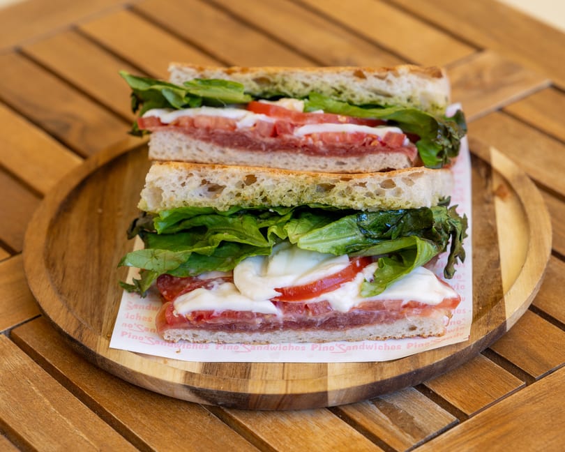 Two halves of a sandwich with pesto, lettuce, tomatoes, and mozzarella on a wooden plate.