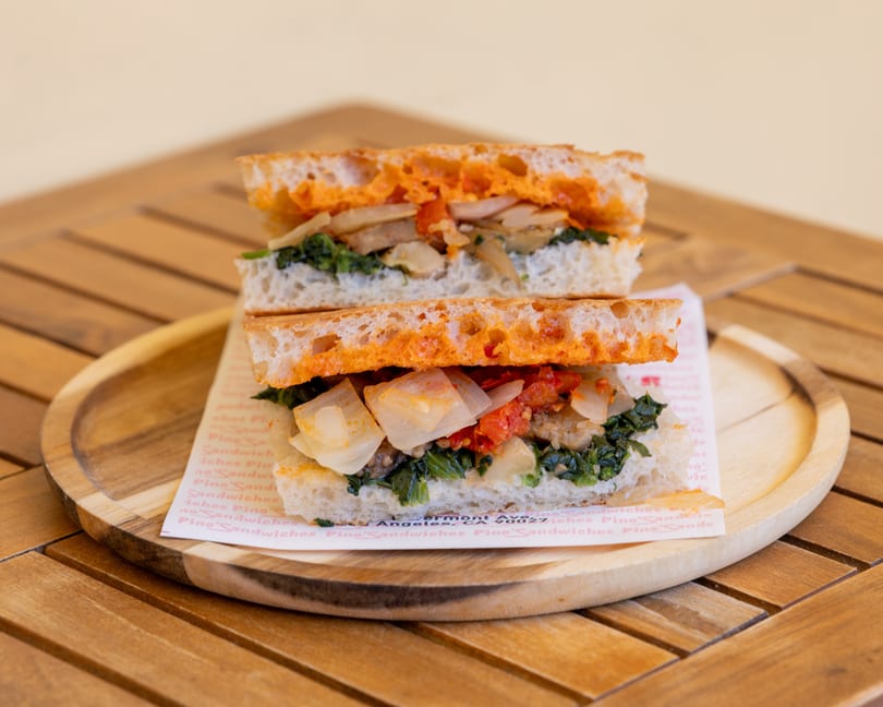 A sliced sandwich with greens, onions, and red sauce on focaccia bread, served on a wooden plate.
